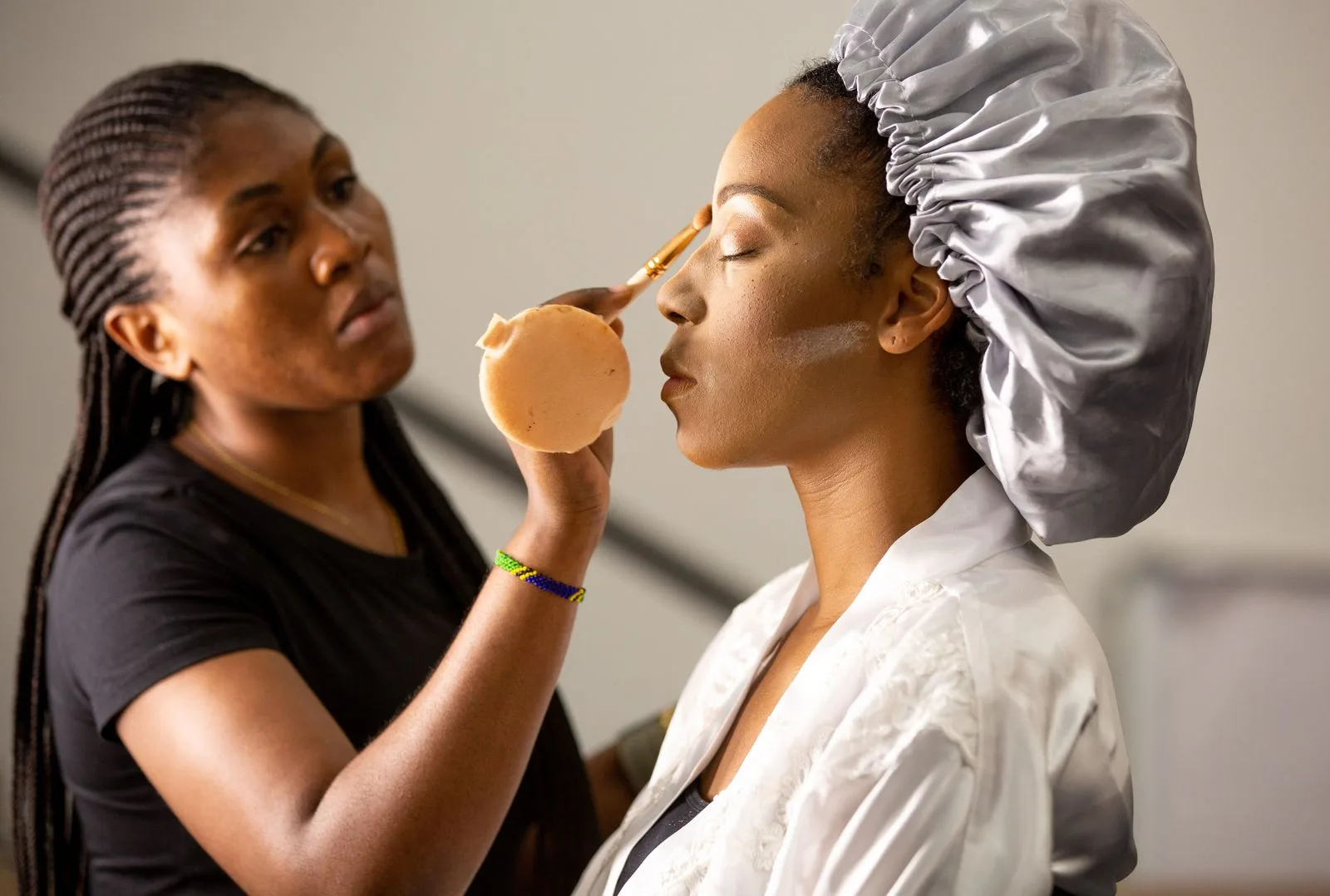 A makeup artist applying foundation to a client in a robe and satin bonnet, the bride tax makeup artist row | Wedded Wonderland